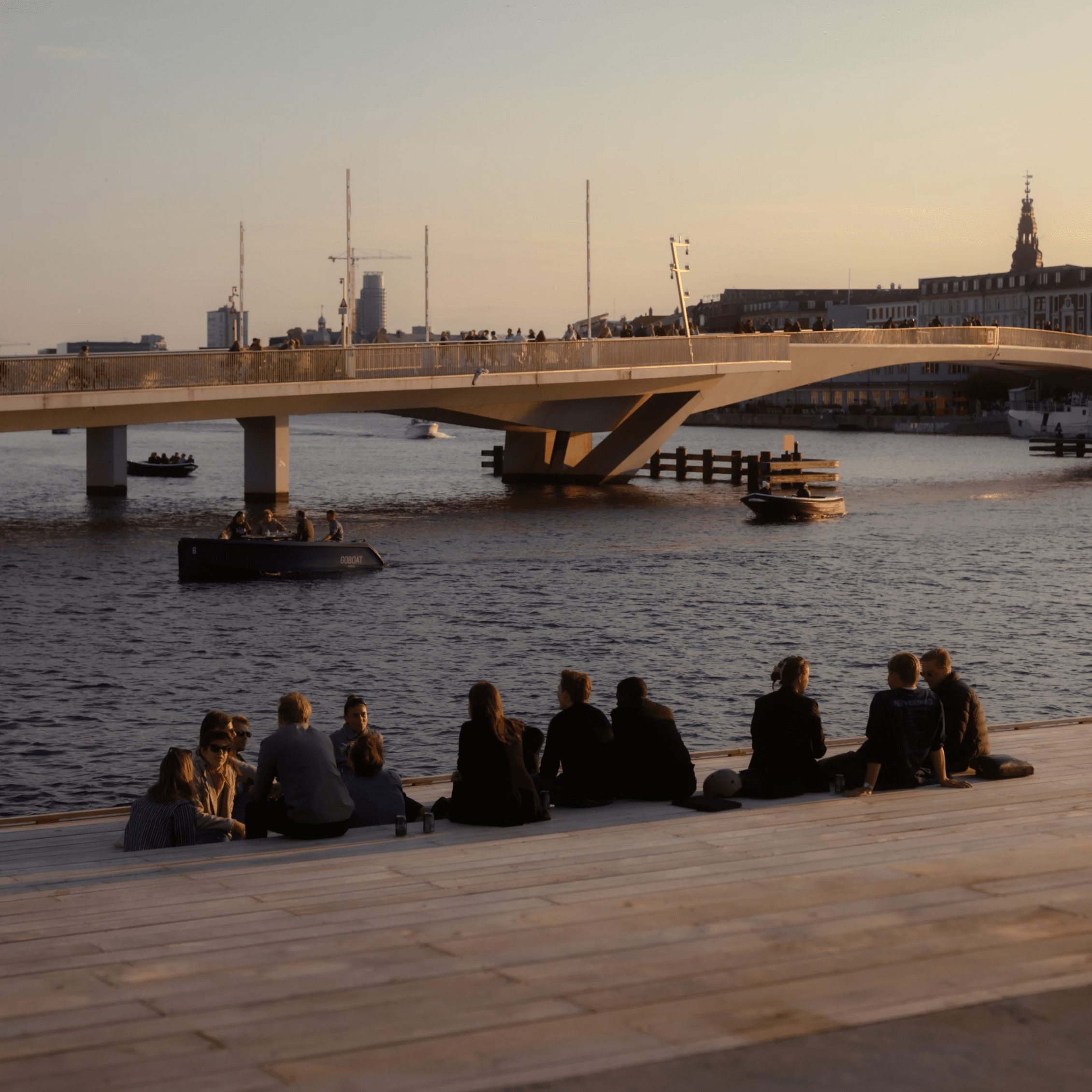 People gathering by the water near a bridge at sunset.
