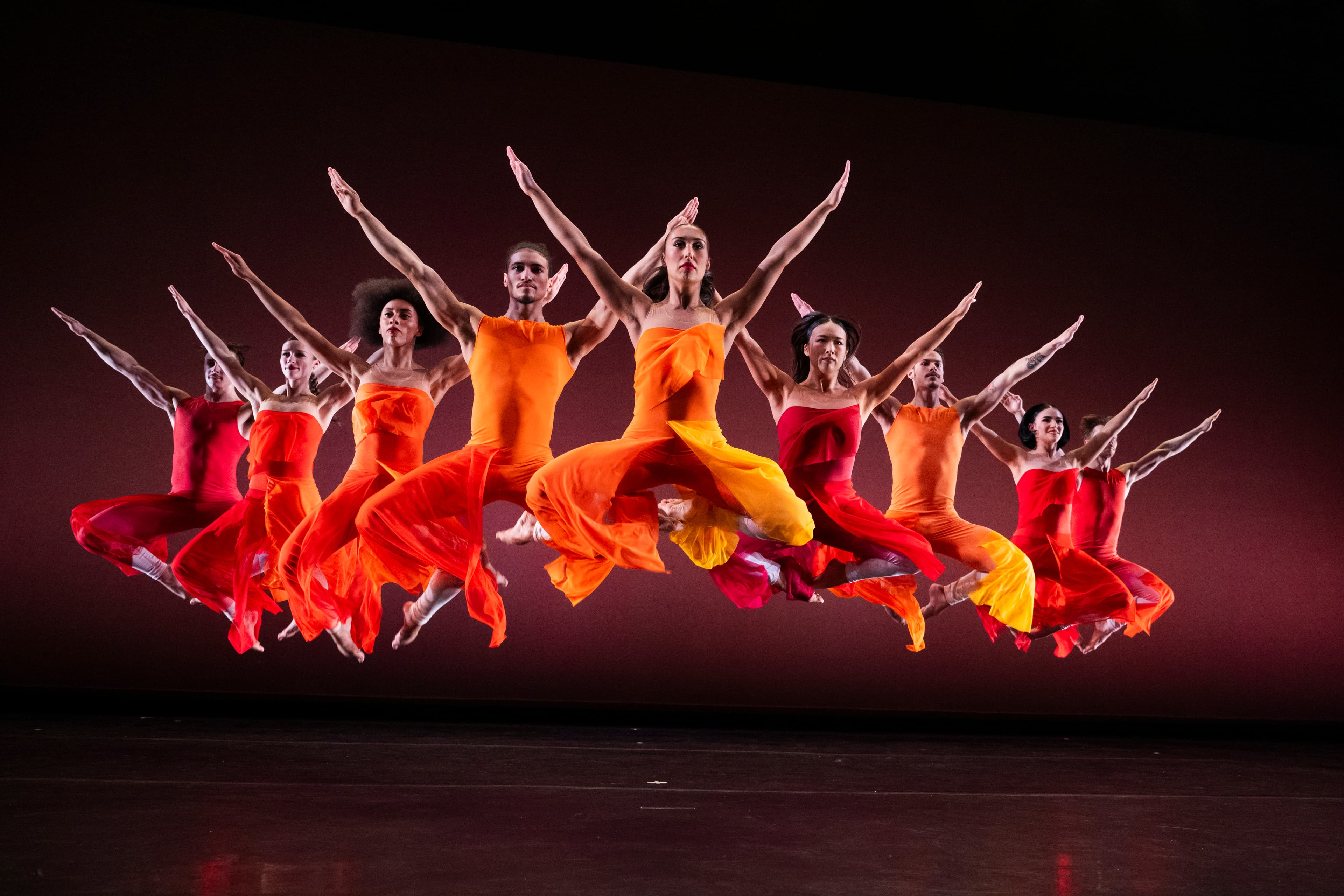 Dancers in red, orange, and yellow costumes leap together onstage with their arms raised.