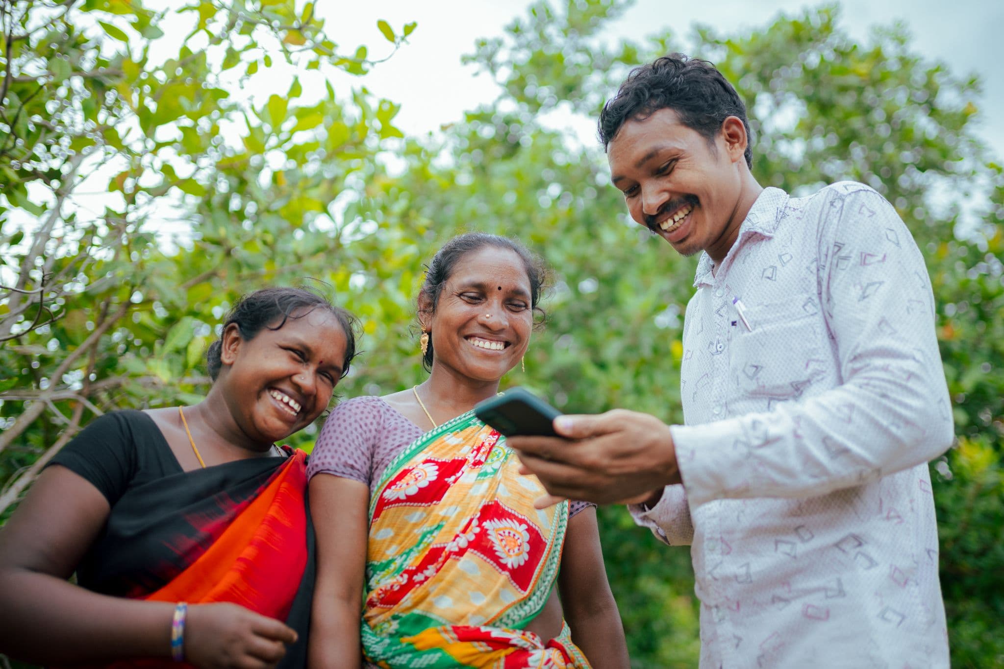 Two women and a man look at a smartphone together outdoors among green trees.