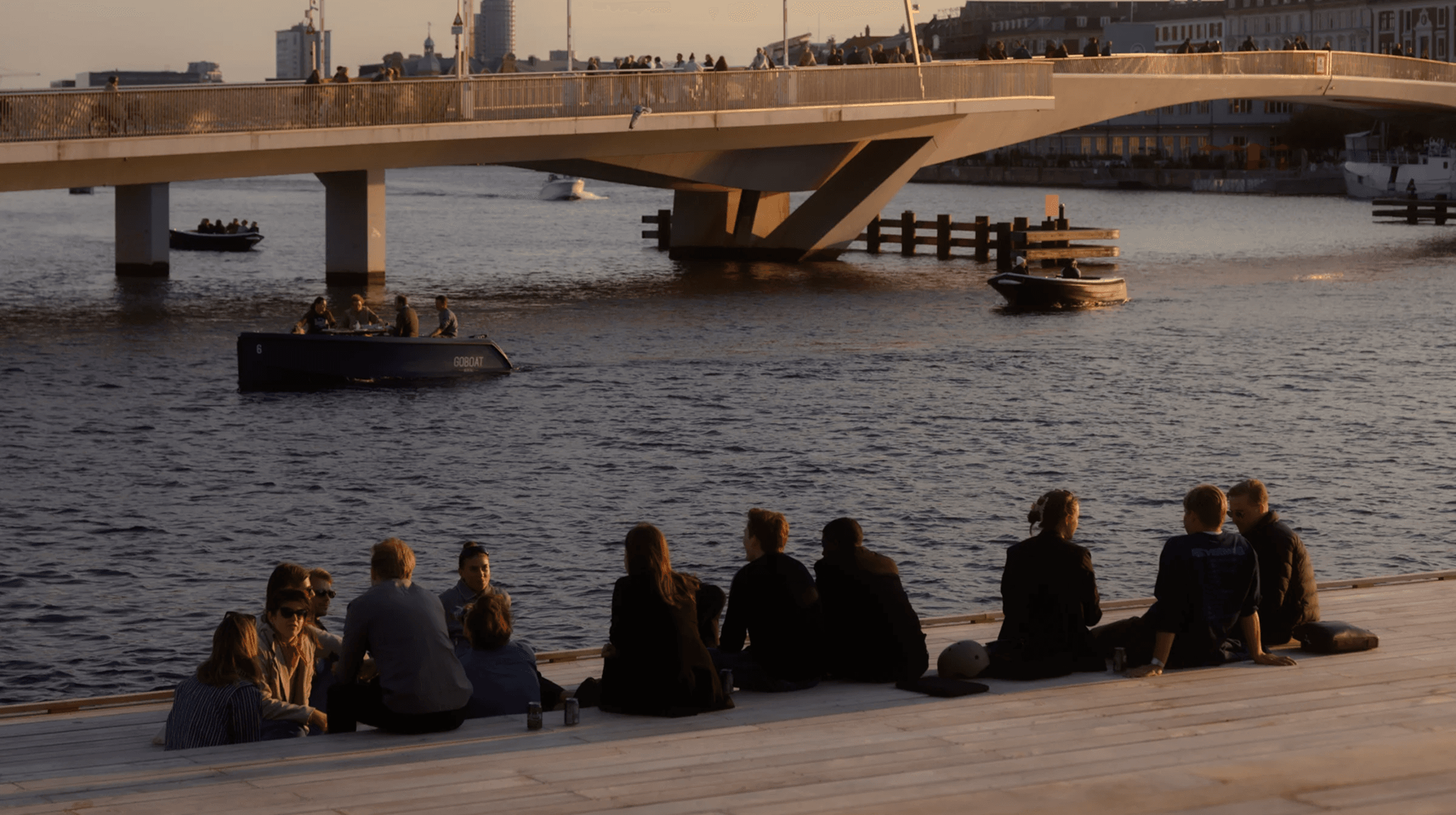 People gathering by the water near a bridge at sunset.